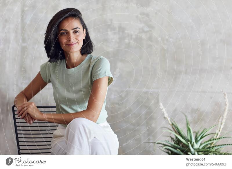 Lächelnde Geschäftsfrau sitzt auf Stuhl gegen graue Wand im Büro Farbaufnahme Farbe Farbfoto Farbphoto Deutschland Innenaufnahme Innenaufnahmen innen drinnen