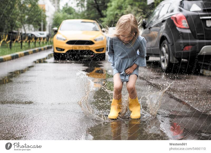 Mädchen in blauem Kleid und Gummistiefeln, springt in Teich auf Straße, gelbes Auto im Hintergrund Pfütze Pfützen Lache Gefahr gefaehrlich Gefahren gefährlich