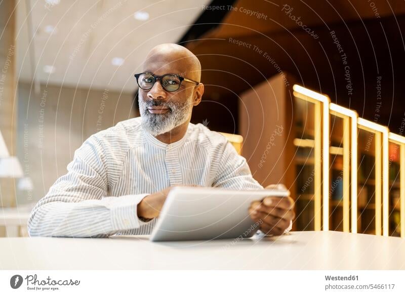 Porträt von reifen Mann sitzt am Schreibtisch in einer Bibliothek mit digitalen Tablette Geschäftsmann Businessmann Businessmänner Geschäftsmänner Portrait
