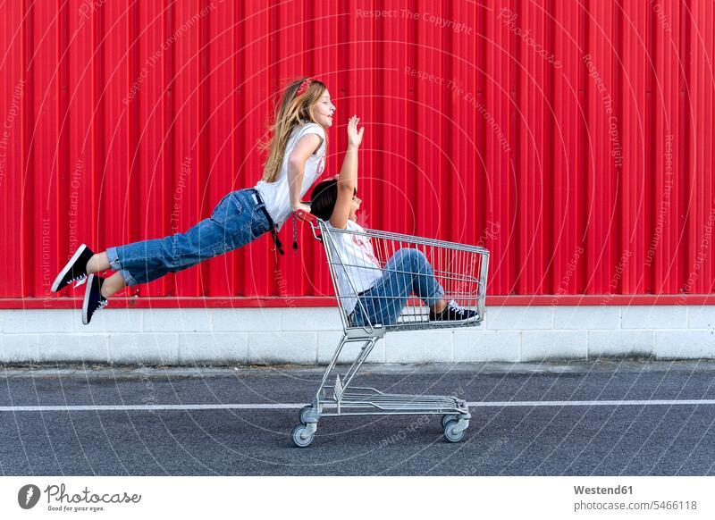 Schwestern mit Einkaufswagen vor roter Wand Hosen Jeanshose T-Shirts Warenkorb Kauf Einkaufen shoppen shopping sitzend sitzt begeistert Enthusiasmus