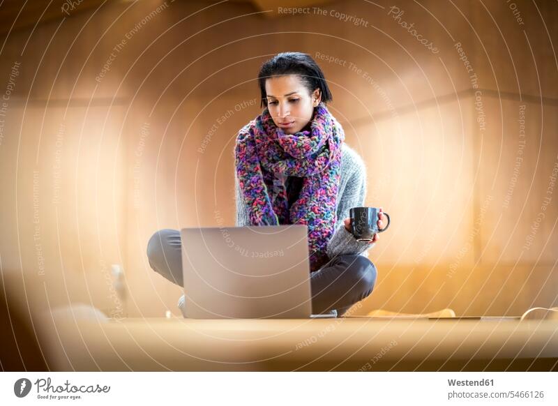Porträt einer modischen jungen Frau mit einer Tasse Kaffee, die mit einem Laptop auf einem Konferenztisch sitzt Notebook Laptops Notebooks benutzen benützen