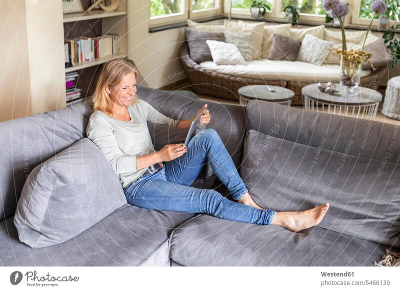 Lachende blonde Frau entspannt sich auf der Couch zu Hause mit digitalem Tablet Leute Menschen People Person Personen Europäisch Kaukasier kaukasisch 1 Ein