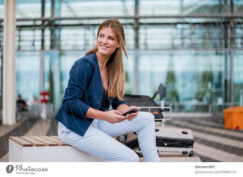 Lächelnde junge Geschäftsfrau sitzt im Freien mit Handy und Koffer und schaut sich um Mobiltelefon Handies Handys Mobiltelefone sitzen sitzend Geschäftsfrauen