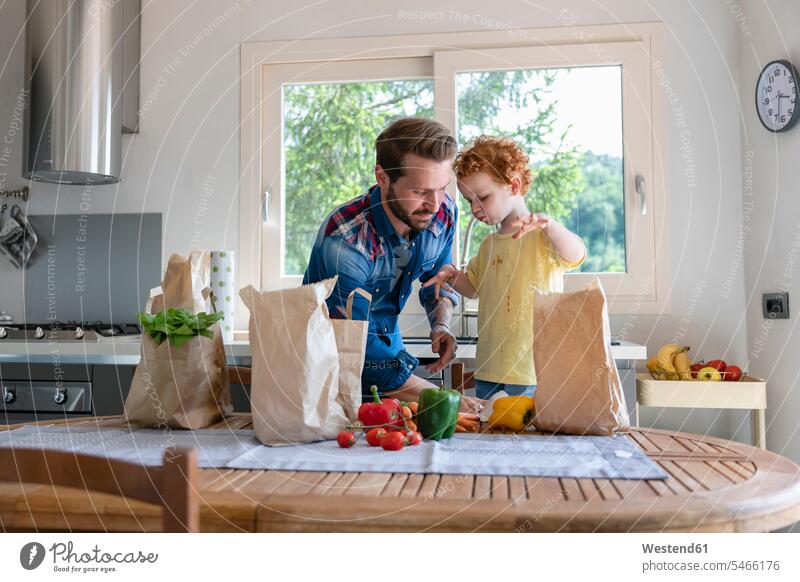 Vater reinigt die Kleidung des Sohnes, während er in der Küche am Esstisch mit Gemüse steht Farbaufnahme Farbe Farbfoto Farbphoto Innenaufnahme Innenaufnahmen