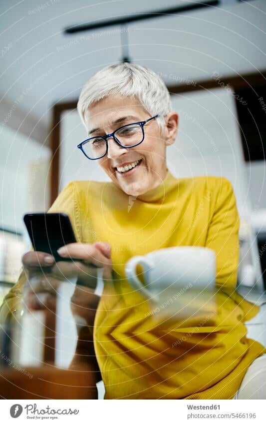 Ältere Frau trinkt Kaffee und überprüft Textnachrichten auf ihrem Smartphone Leute Menschen People Person Personen Europäisch Kaukasier kaukasisch erwachsen