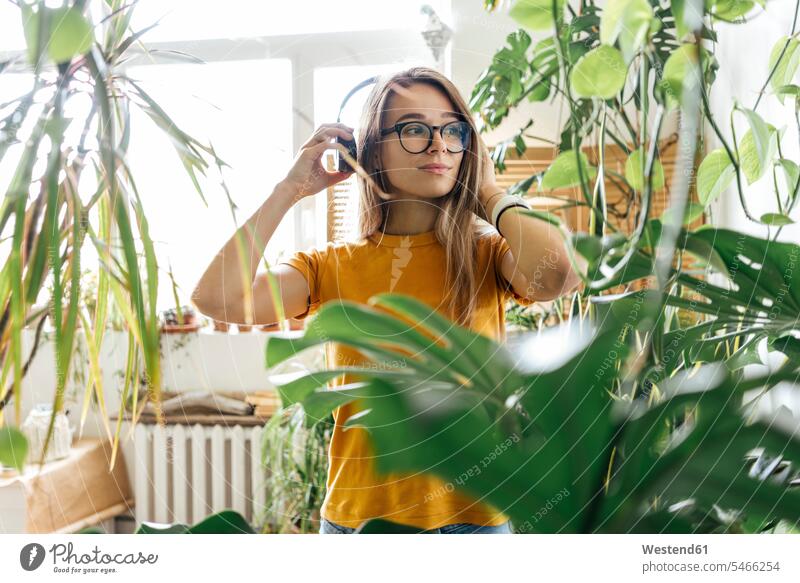 Junge Frau, umgeben von Pflanzen, die mit Kopfhörern Musik hört T-Shirts Kopfhoerer Brillen hoeren geniessen Genuss zufrieden Farben Farbtoene Farbton Farbtöne