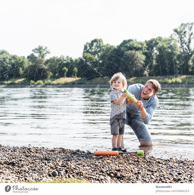 Vater und Sohn haben Spaß am Flussufer, spielen mit einer Wasserpistole lachen Wasserkanone Wasserspritze Papas Väter Vati Vatis Papis Söhne Sommer Sommerzeit