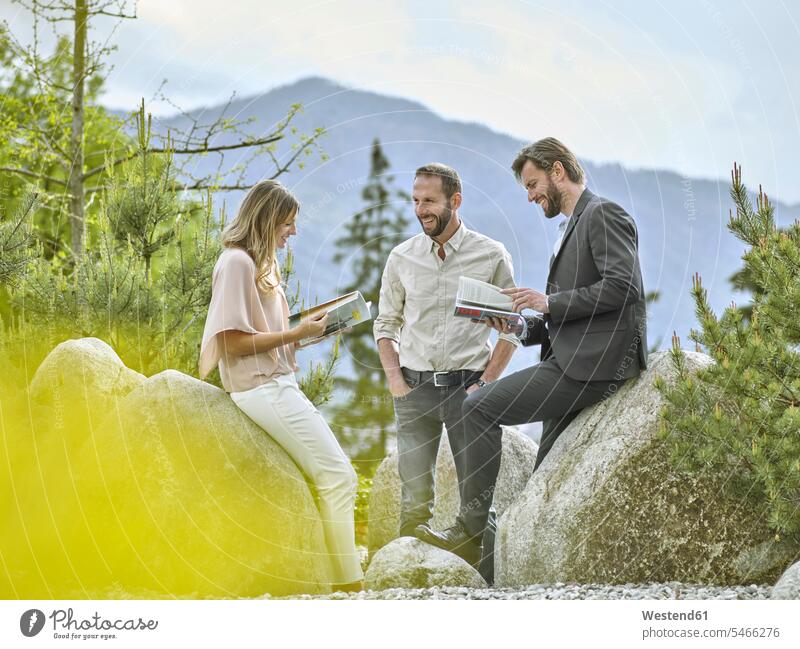 Lächelnde Kollegen mit Zeitschriften, die eine Pause an Felsen machen lächeln sprechen Gestein Magazin Beruf ländliches Motiv Baum Teamwork Zusammenarbeit Tirol