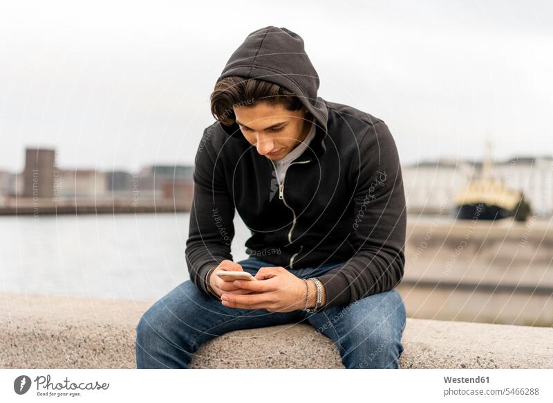 Dänemark, Kopenhagen, junger Mann sitzt auf der Mauer am Wasser und benutzt ein Mobiltelefon Kobenhavn chatten Chat Smartphone iPhone Smartphones Reisende