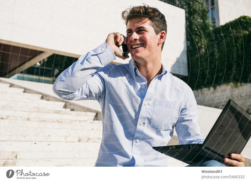 Lächelnder junger Mann, der an einem sonnigen Tag mit einem Laptop sitzt und telefoniert Farbaufnahme Farbe Farbfoto Farbphoto Spanien Außenaufnahme außen