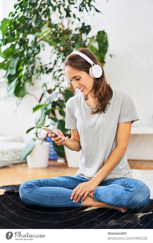 Lächelnde junge Frau sitzt auf dem Boden mit Kopfhörern und Handy Kopfhoerer Mobiltelefon Handies Handys Mobiltelefone sitzen sitzend weiblich Frauen Zuhause