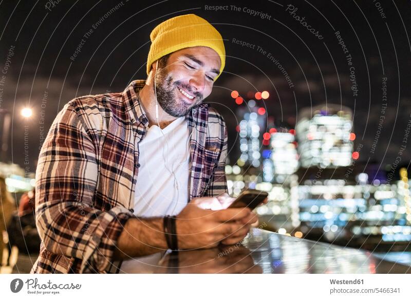 UK, London, lächelnder Mann lehnt an einem Geländer und schaut auf sein Telefon mit Stadt Lichter im Hintergrund Männer männlich Nacht nachts ansehen Handy