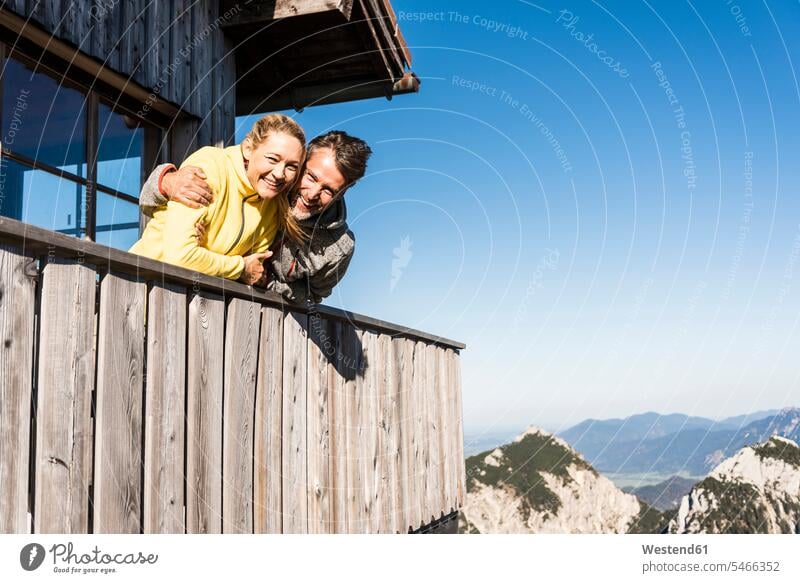 Paar lehnt auf Balkon einer Berghütte, mit Armen um Arm umlegen Arme umlegen Gebirge Berglandschaft Gebirgslandschaft Gebirgskette Gebirgszug Berge aufstützen