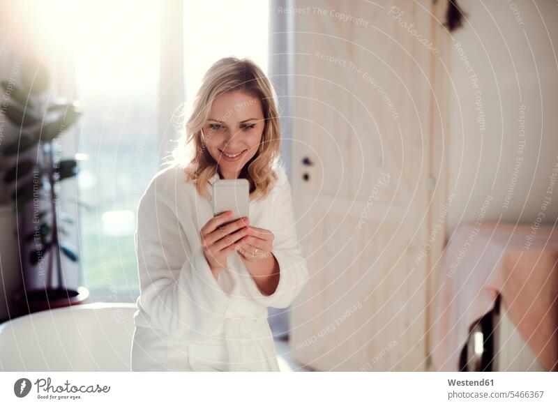 Lächelnde Frau im Bademantel im Bad zu Hause hält Handy Zuhause daheim weiblich Frauen Badezimmer morgens Morgen früh Frühe lächeln halten Mobiltelefon Handies