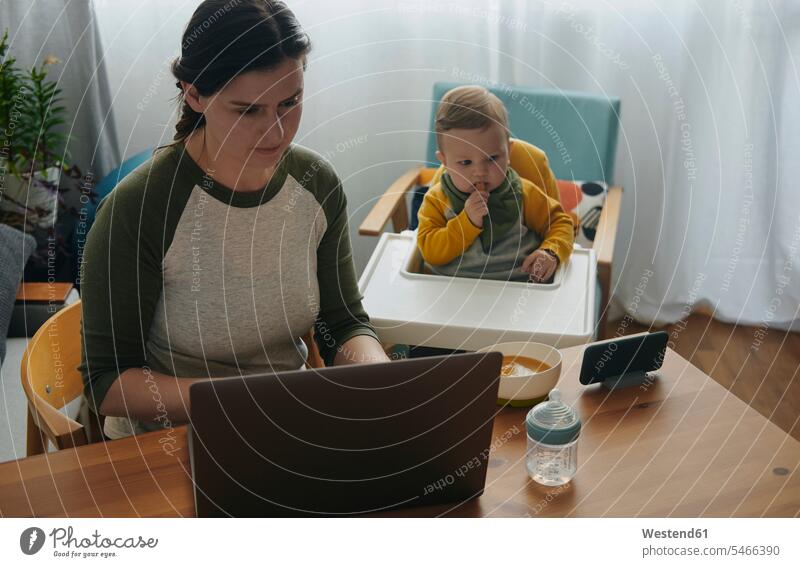 Frau arbeitet zu Hause am Laptop, während ihr kleiner Sohn Filme auf dem Handy anschaut Leute Menschen People Person Personen Europäisch Kaukasier kaukasisch 2