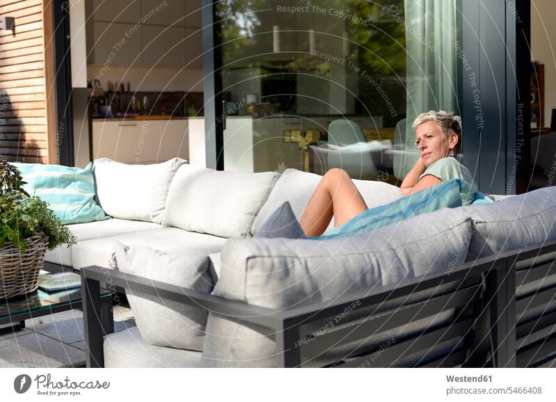 Nachdenkliche Frau sitzt auf der Couch auf der Terrasse ihres Hauses weiblich Frauen Sofa Couches Liege Sofas Terrassen sitzen sitzend nachdenklich Häuser