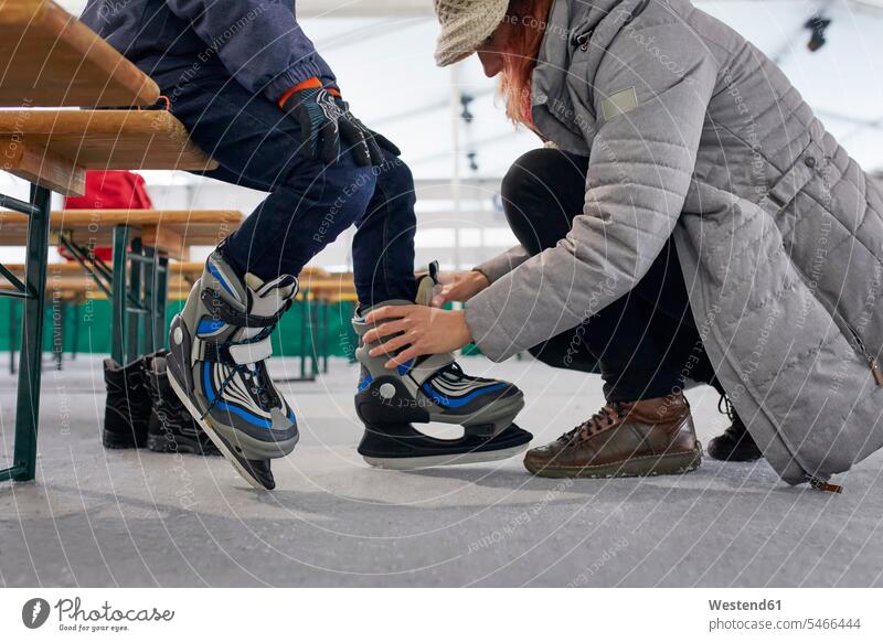 Mutter hilft ihrem Sohn, Schlittschuhe anzuziehen helfen mithelfen Hilfsbereitschaft beistehen Mithilfe Beistand Hilfe Söhne Eisbahn Eislaufbahn