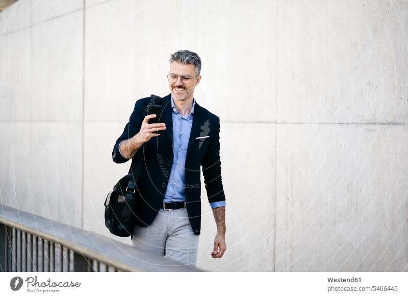 Lächelnder grauhaariger Geschäftsmann geht mit seinem Handy durch die Stadt geschäftlich Geschäftsleben Geschäftswelt Geschäftsperson Geschäftspersonen