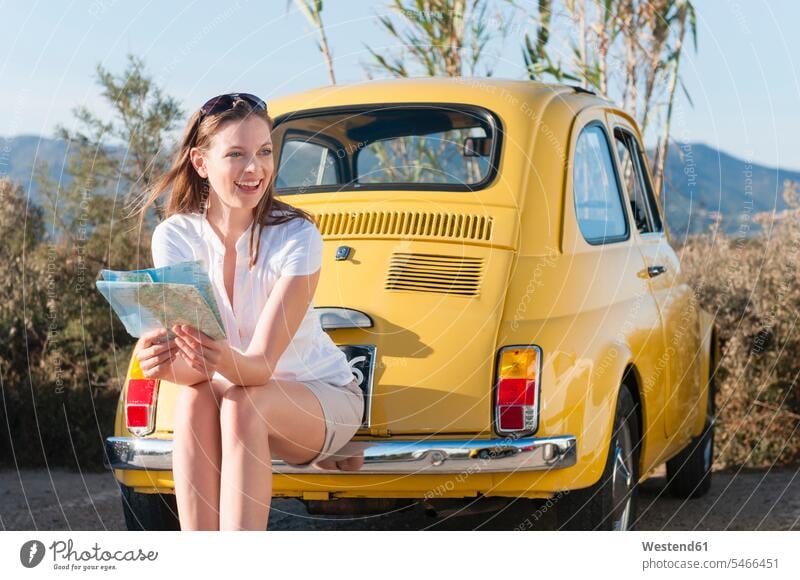 Porträt einer lachenden Frau mit Karte, die auf der Stoßstange eines gelben Oldtimers sitzt, Sardinien, Italien Karten Landkarten KFZ Verkehrsmittel Automobil