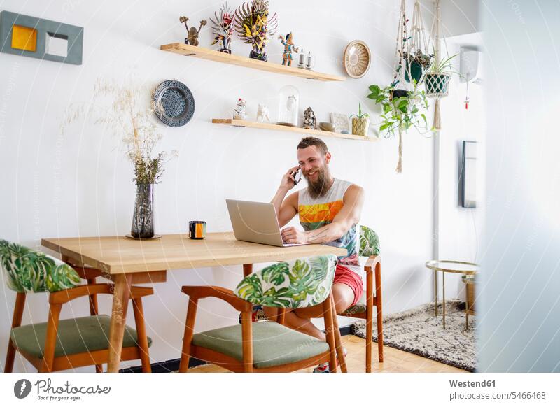 Mittelgroßer erwachsener Mann spricht am Smartphone, während er im Wohnzimmer am Laptop sitzt Farbaufnahme Farbe Farbfoto Farbphoto Innenaufnahme Innenaufnahmen