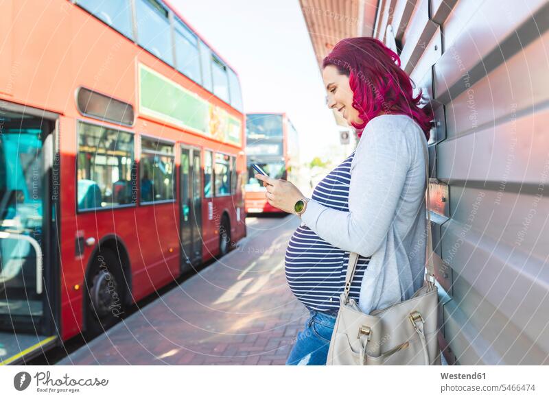 Schwangere Frau benutzt ihr Telefon an der Bushaltestelle Oberkoerper Oberkörper Torso Torsos Bäuche Transport Transportwesen KFZ Verkehrsmittel Autobus