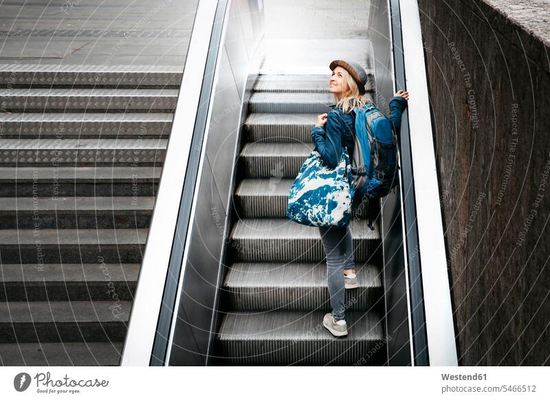 Frau mit Rucksack und Reisetasche steht auf Rolltreppe und schaut nach oben Europäer Kaukasier Europäisch kaukasisch Innenaufnahme drinnen Innenaufnahmen