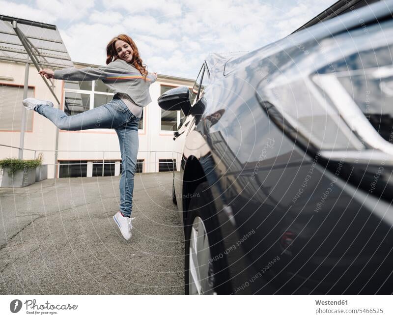 Glückliche rothaarige Frau springt neben Auto KFZ Verkehrsmittel Automobil Autos PKW PKWs Wagen begeistert Enthusiasmus enthusiastisch Überschwang