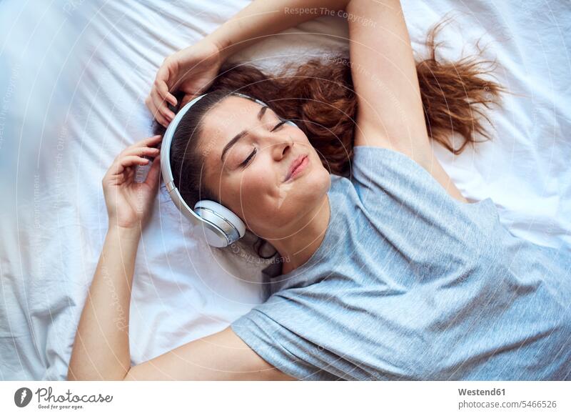 Porträt einer glücklichen jungen Frau mit geschlossenen Augen, die auf dem Bett liegt und mit Kopfhörern Musik hört Leute Menschen People Person Personen