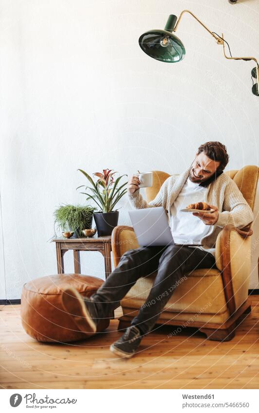 Lächelnder Mann am Telefon, der zu Hause auf einem Sessel mit Laptop, Croissant und einer Tasse Kaffee sitzt Leute Menschen People Person Personen Europäisch