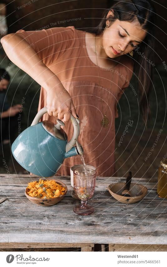 Schöne Frau gießt heißes Wasser in Glas, während sie Kräutertee auf Holztisch zubereitet Farbaufnahme Farbe Farbfoto Farbphoto Innenaufnahme Innenaufnahmen