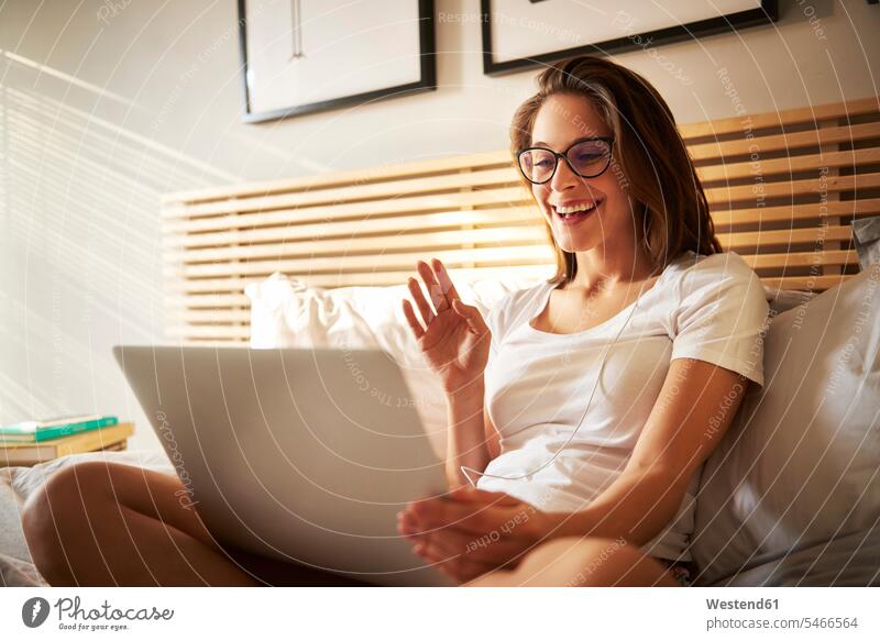 Porträt einer lachenden jungen Frau, die mit einem Laptop auf dem Bett sitzt und plaudert sitzen sitzend benutzen benützen Betten weiblich Frauen Portrait
