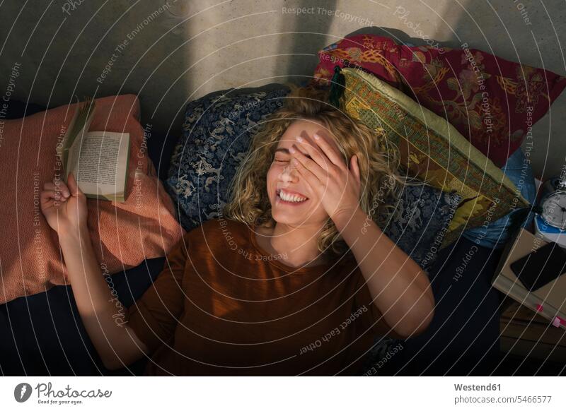 Porträt einer lachenden jungen Frau, die auf dem Bett liegt und ein Buch liest Leute Menschen People Person Personen Europäisch Kaukasier kaukasisch 1 Ein