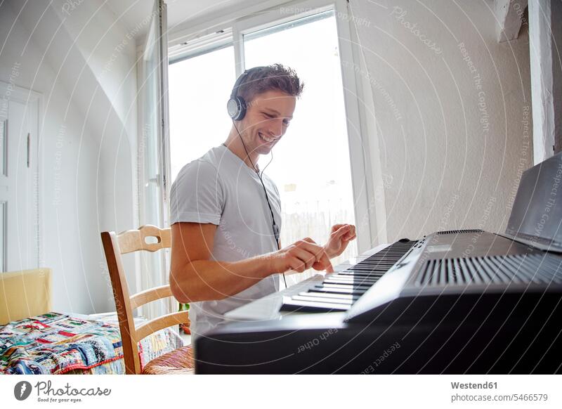 Lächelnder junger Mann zu Hause mit Kopfhörern spielt Digitalpiano Kopfhoerer Klavier Piano Pianos Klaviere Zuhause daheim Männer männlich spielen