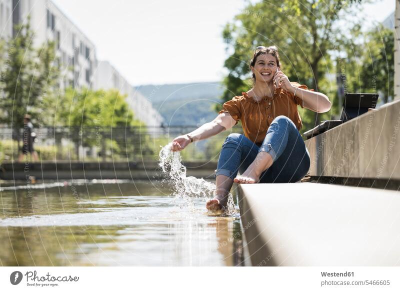 Lächelnde Frau spielt mit Teichwasser, während sie im Park über ein Smartphone telefoniert Farbaufnahme Farbe Farbfoto Farbphoto Deutschland