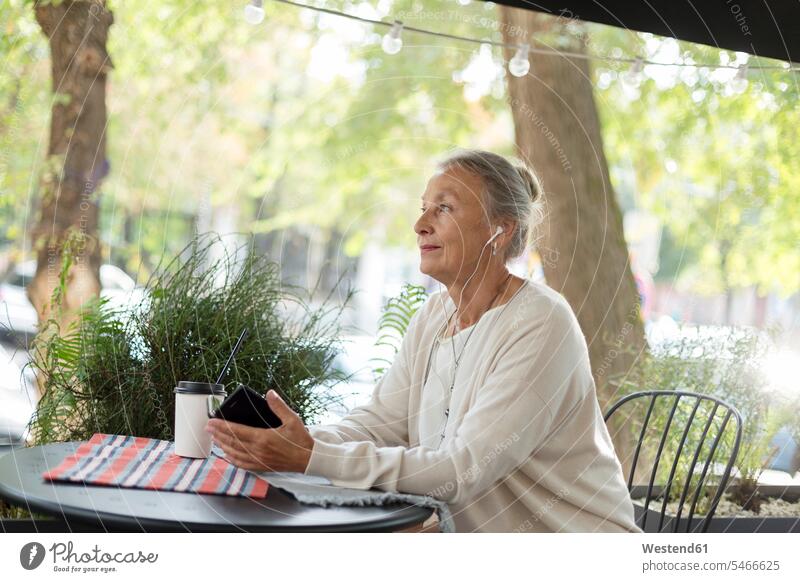 Ältere Frau in einem Straßencafé mit Handy und Kopfhörern Straßencafe Straßencafes Strassencafe Strassencafes Ohrhörer weiblich Frauen Seniorin älter