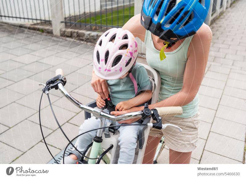 Mutter und Tochter fahren Fahrrad, Tochter mit Helm sitzt im Kindersitz und schnallt sich an Helme Mami Mutti Mütter Mama Töchter anschnallen sitzen sitzend