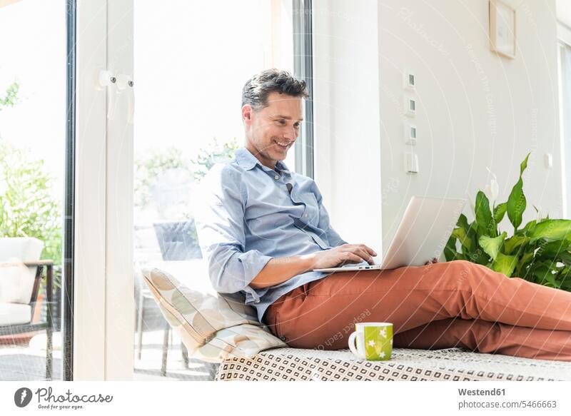 Reifer Mann sitzt auf dem Sofa und benutzt einen Laptop Rechner Laptops Notebook Notebooks sitzend freuen Frohsinn Fröhlichkeit Heiterkeit Glück glücklich sein