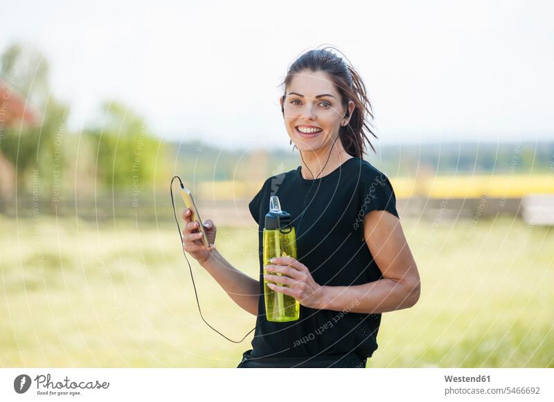 Sportliche Frau mit Smartphone in Kühlpause Musik hören lächeln telefonieren anrufen Anruf telephonieren Ohrhörer iPhone Smartphones Wasserflasche Flasche