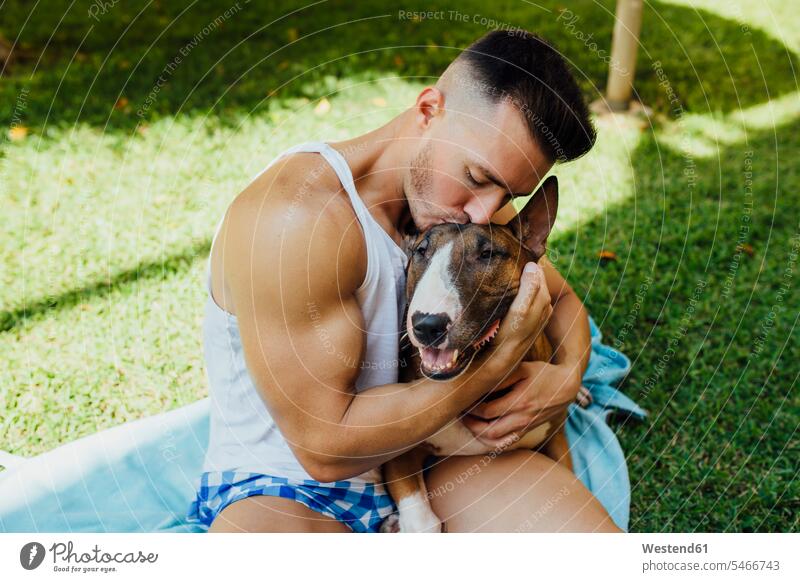 Muskulöser Mann sitzt auf einer Decke auf einer Wiese und umarmt seinen Hund Tiere Tierwelt Haustiere Hunde Decken entspannen relaxen sitzend Arm umlegen