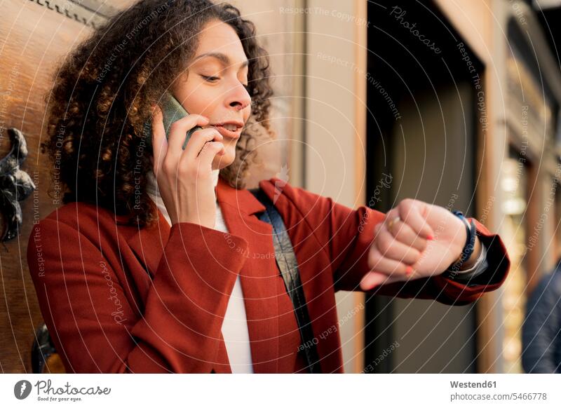 Frau am Telefon in der Stadt, die die Uhrzeit überprüft Leute Menschen People Person Personen gelockt gelockte Haare gelocktes Haar lockig lockiges Haar Jacken