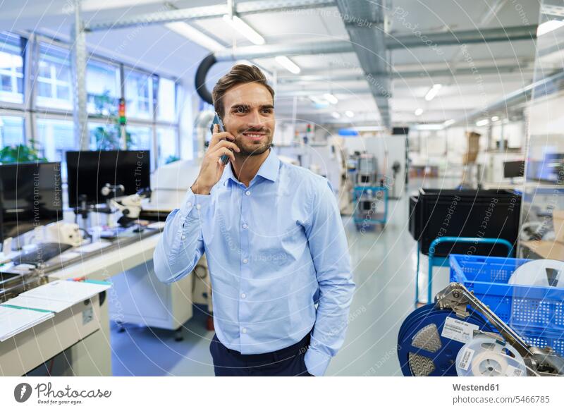 Lächelnder junger männlicher Berufstätiger spricht am Smartphone, während er in der beleuchteten Fabrik steht Farbaufnahme Farbe Farbfoto Farbphoto