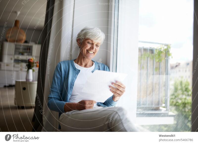 Ältere Frau liest fröhlich Brief, während sie zu Hause am Fenster sitzt Farbaufnahme Farbe Farbfoto Farbphoto Innenaufnahme Innenaufnahmen innen drinnen Tag