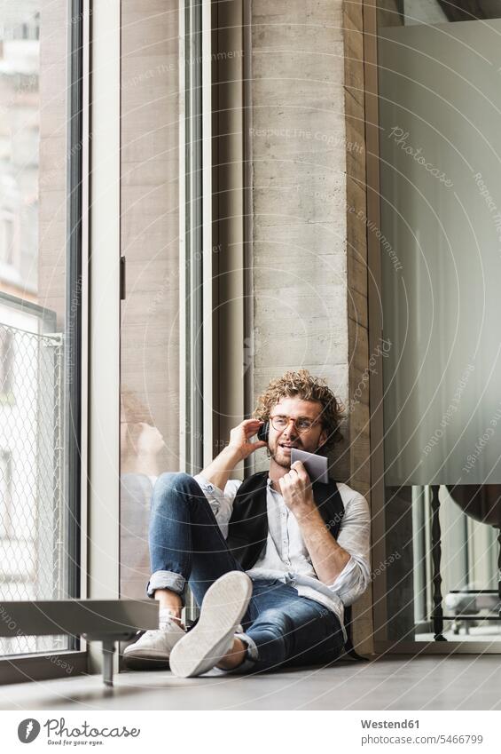 Lässiger junger Mann, der mit Kopfhörern und Phablet am Fenster sitzt Männer männlich Smartlet Handy Mobiltelefon Handies Handys Mobiltelefone Kopfhoerer leger