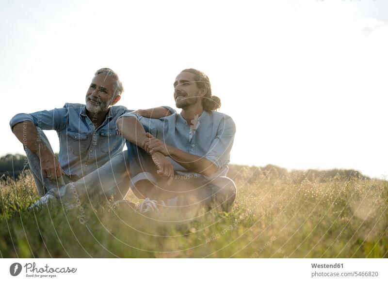 Erwachsener Vater mit erwachsenem Sohn auf einer Wiese auf dem Land sitzend Hemden entspannen relaxen sitzt zufrieden auf dem Lande Muße Bindung Gemeinschaft
