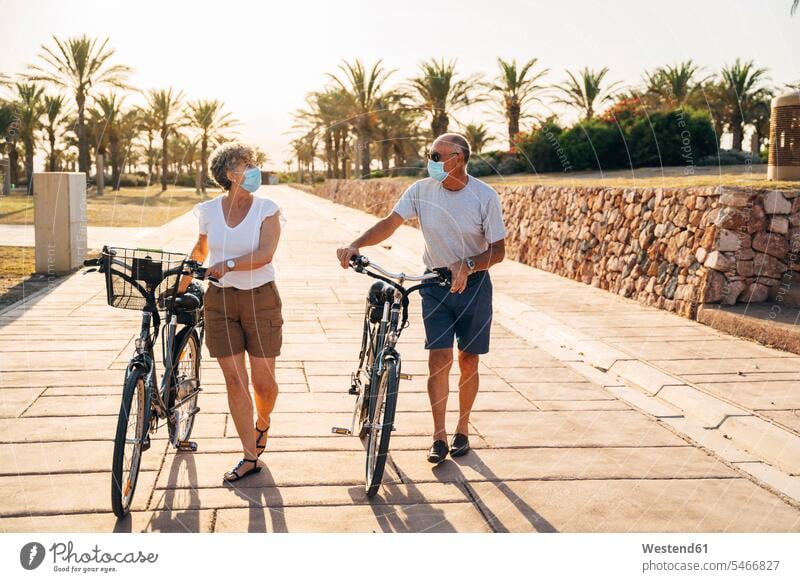 Älterer Mann und Frau mit Gesichtsmaske unterhalten sich beim Gehen mit dem Fahrrad im Park Farbaufnahme Farbe Farbfoto Farbphoto Außenaufnahme außen draußen