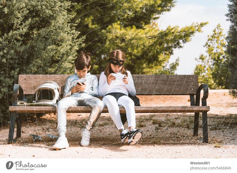 Zwei Kinder in Astronauten- und Superheldenkostümen, die ihr Handy auf der Parkbank benutzen Freunde Kameradschaft Leute Menschen People Person Personen Held