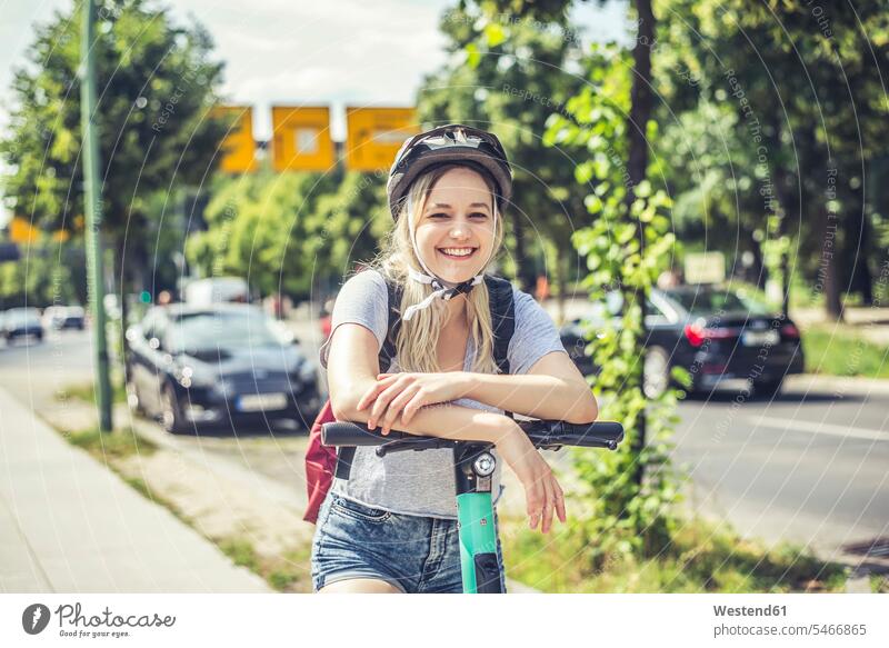 Porträt einer glücklichen jungen Frau mit Fahrradhelm, die sich an den Lenker eines E-Scooters lehnt Rucksäcke KFZ Verkehrsmittel Automobil Autos PKW PKWs Wagen