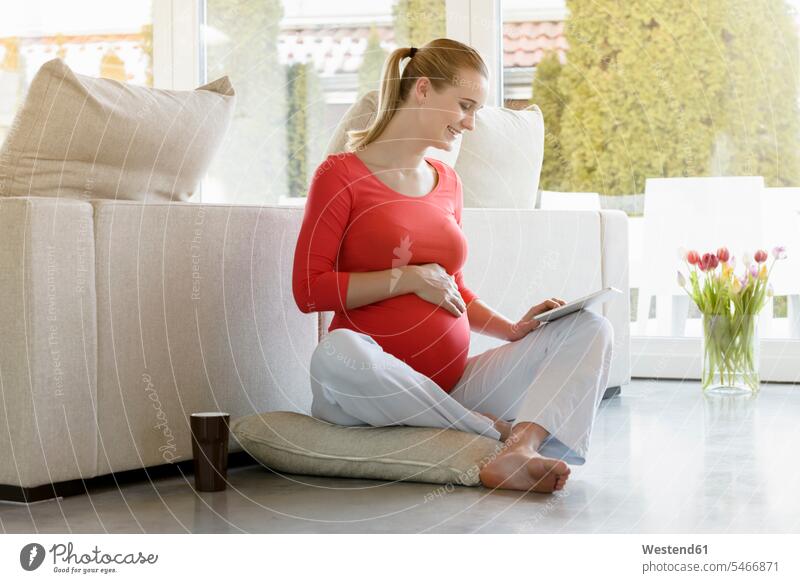 Lächelnde schwangere Frau sitzt zu Hause auf dem Boden und benutzt Tablette Schwangere Böden Boeden lächeln weiblich Frauen sitzen sitzend Zuhause daheim