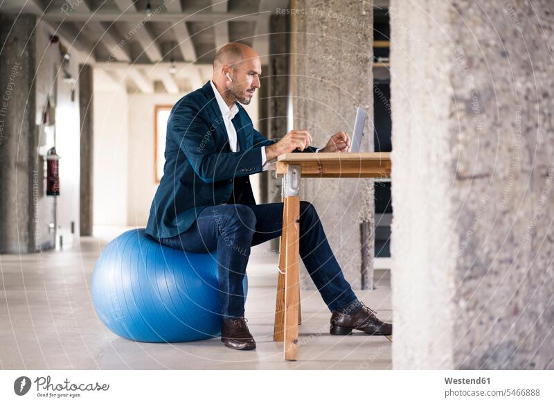 Mann benutzt Laptop, während er im Büro auf einem Fitnessball sitzt Farbaufnahme Farbe Farbfoto Farbphoto Innenaufnahme Innenaufnahmen innen drinnen Tag