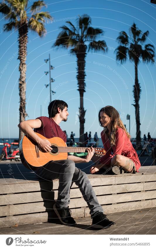 Spanien, Barcelona, lächelndes Paar mit einer Gitarre auf einer Bank an der Strandpromenade sitzend Sitzbänke Bänke Sitzbank Gitarren Promenade Promenaden